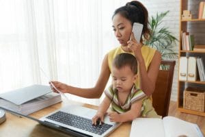 a mom works in front of a computer, on the phone with a document in her hand and a baby on her lap